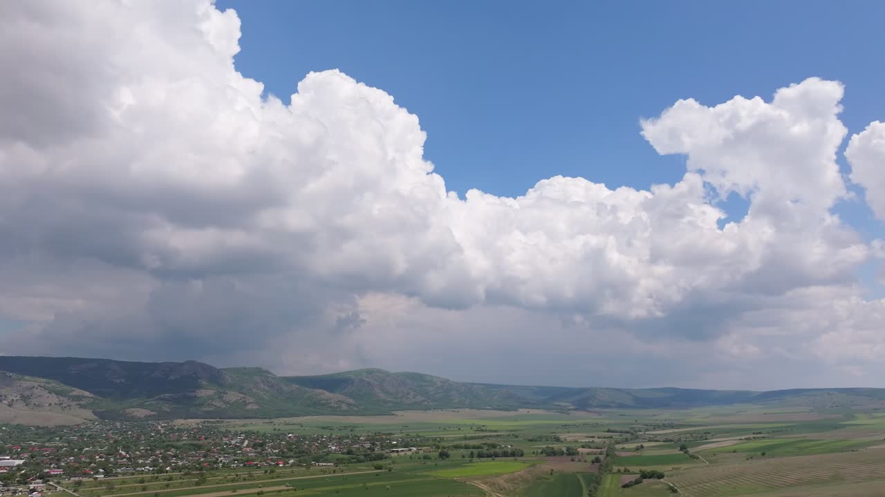 Rural landscape framed by dramatic cumulus clouds and layered mountains stretching into the horizon. Wide panoramic view of green farmlands, a small settlement with red rooftops