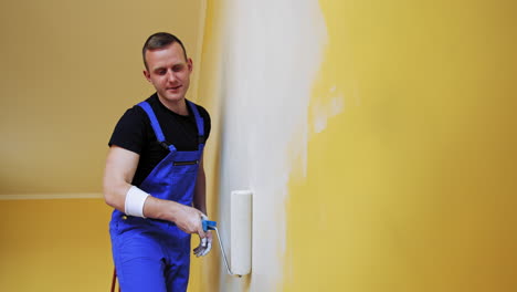 Portrait of a painter in action. Young man builder in blue overalls decorating room with a paint roller in the apartment. Room makeover.
