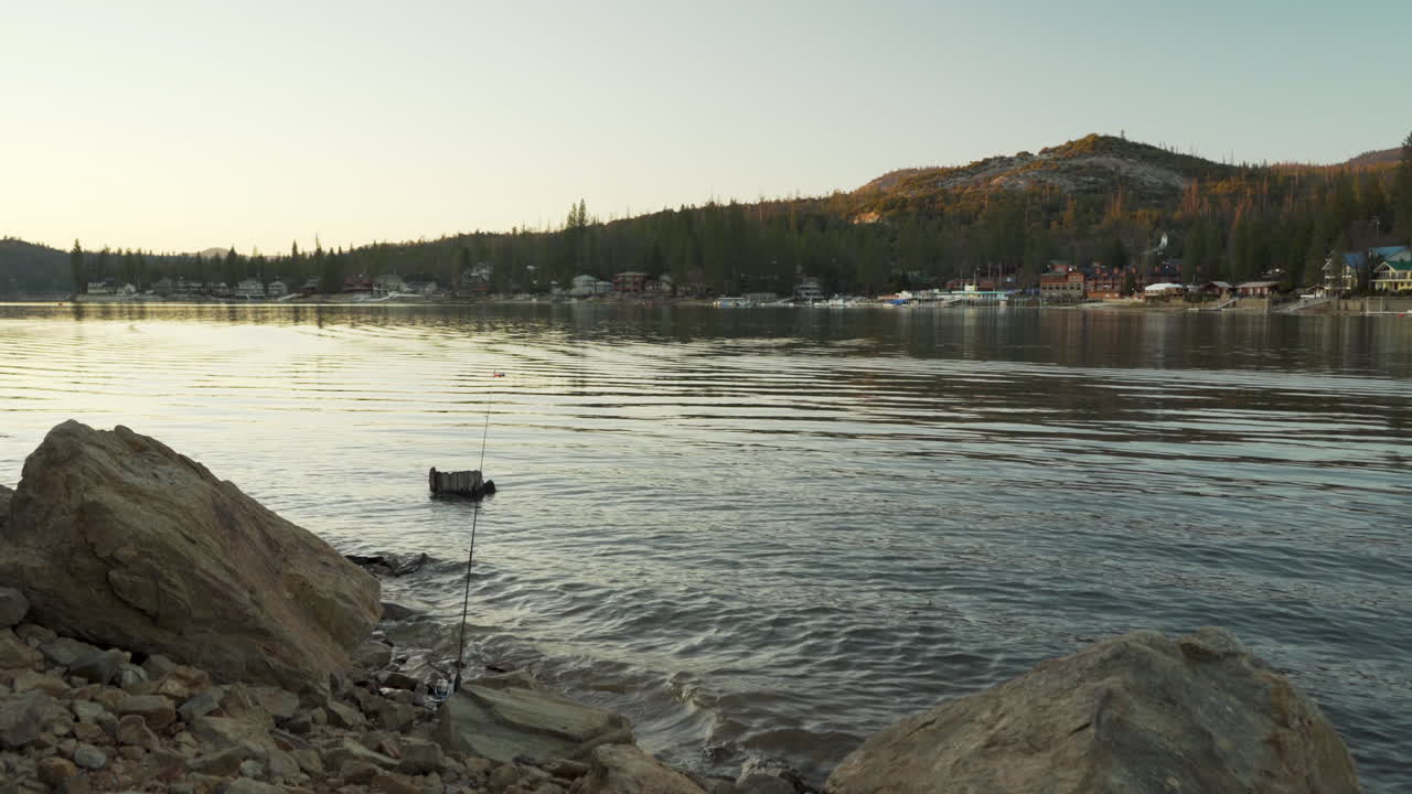 해질녘에 산 호수의 해안선에 있는 바위 사이에 낚싯대가 끼어 있고, 배경에는 고급 주택이 있습니다.