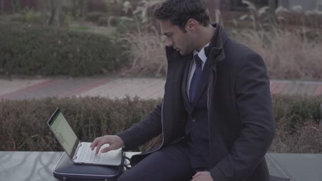 business man outside work one hand on laptop in suit sitiing on the bench