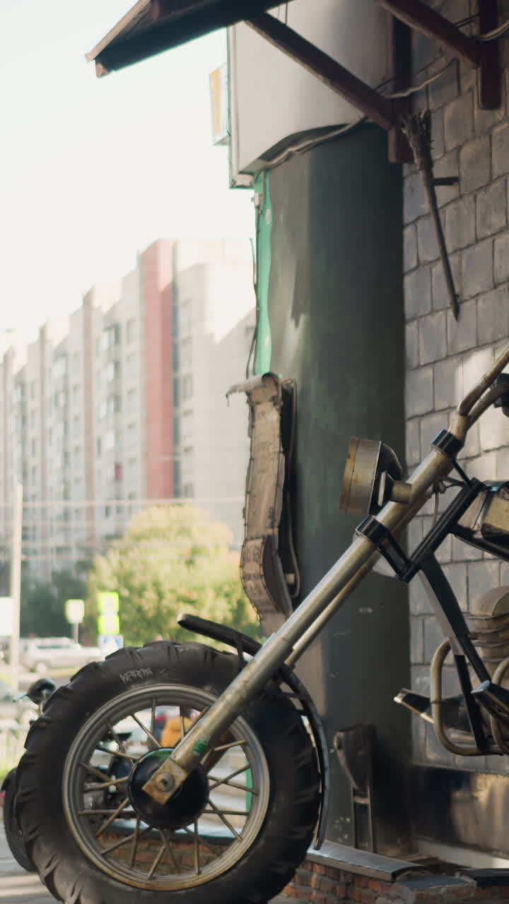 흐릿한 햇빛 아래 도시 거리의 풍화된 기둥에 기대어 있는 빈티지 오토바이 앞바퀴 클로즈업 사진입니다. 크롬 포크와 마모된 타이어에는 녹과 벗겨진 페인트가 보이고, 아파트 건물은 배경으로 흐릿하게 보입니다.