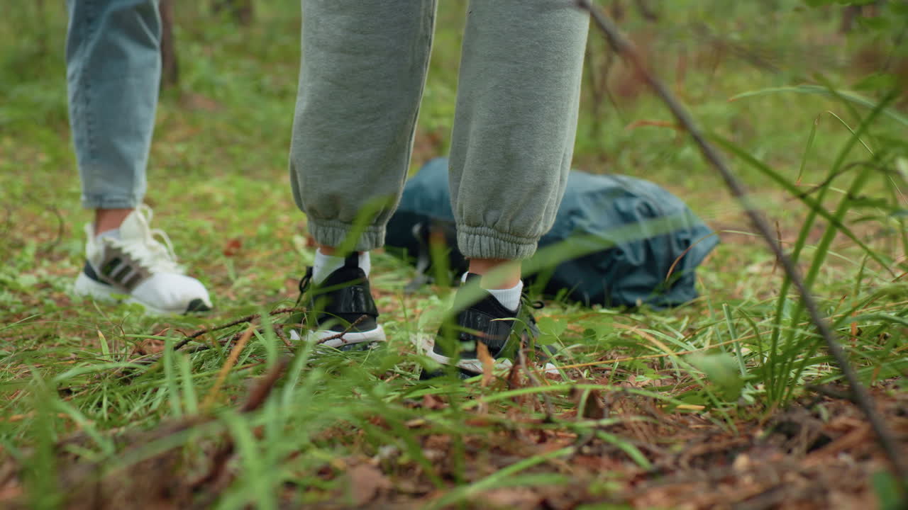 Leg view of people standing in lush forest wearing sneakers and sweatpants as they place travel backpack on grassy forest floor, surrounded by green vegetation, preparing to rest or explore outdoors