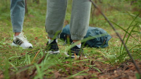 leg view of people standing in lush forest wearing sneakers and sweatpants as they place travel backpack on grassy forest floor, surrounded by green vegetation, preparing to rest or explore outdoors