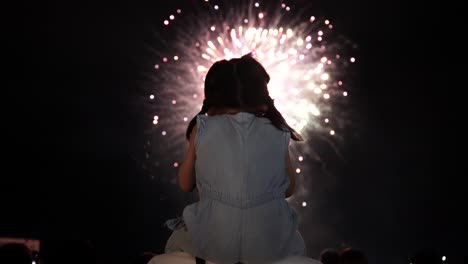 a young asian girl is seen on the back, watching fireworks explode in the sky as she is sitting on her father's shoulders.