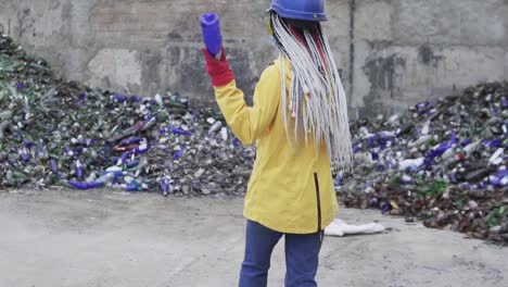 Mujer-Con-Casco-Parada-Contra-El-Montón-De-Vidrios-Rotos,-Botellas-Usadas-Al-Lado-De-La-Pared.-Chica-Con-Chaqueta-Amarilla-Rompiendo-Viejas-Botellas-De-Vidrio-Para-Su-Posterior-Reciclaje.-Vista-Rara