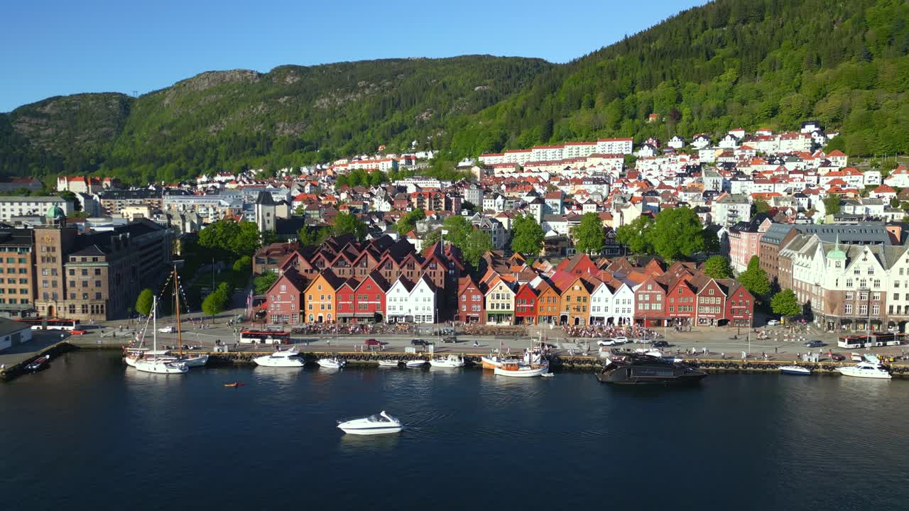 High-altitude aerial view of Bryggen wharf in Bergen, Norway, revealing its colorful wooden buildings along the waterfront, framed by deep blue harbor waters and towering green mountains.