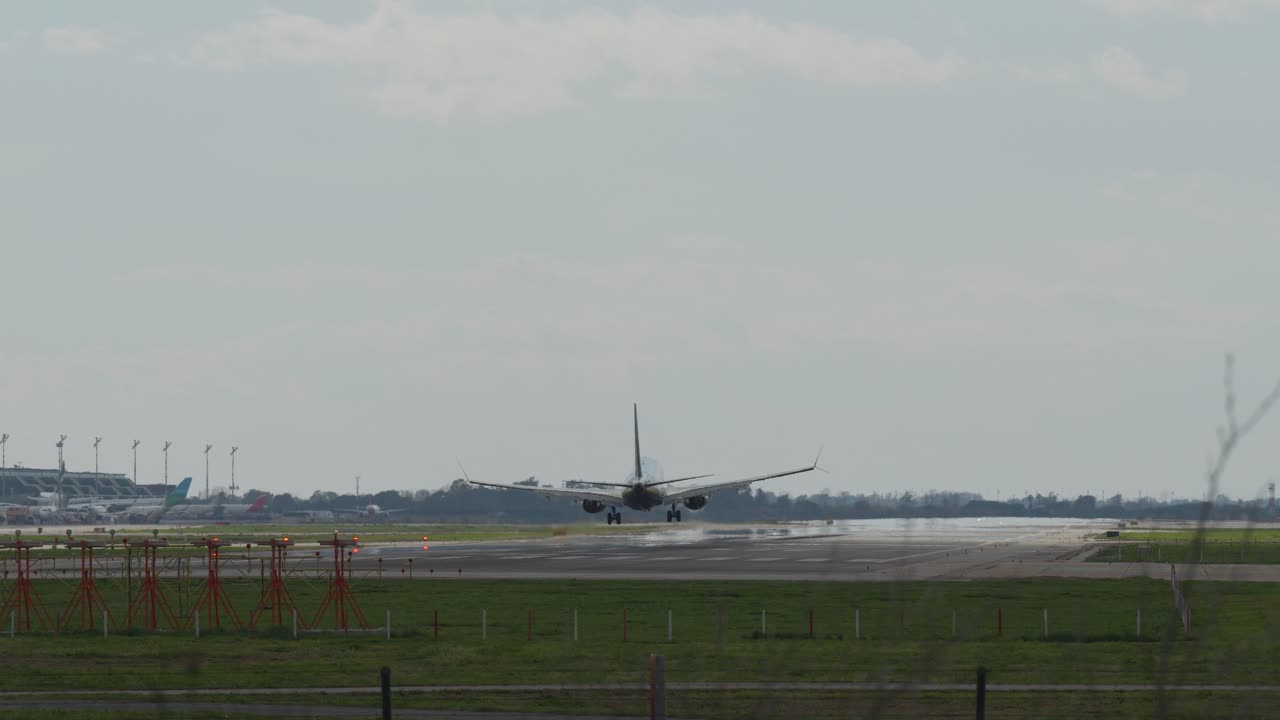 Passenger aircraft approaching runway, descending over illuminated airport infrastructure and green landscape with security monitoring cameras against cloudy sky backdrop