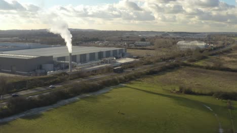 rising aerial drone shot of factory with smoking chimney over fields and motorway