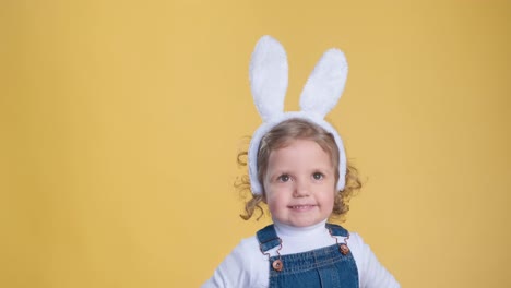 a happy blond toddler with bunny ears and overalls, wearing a smile on her face, can be seen in a joyful gesture on a yellow background