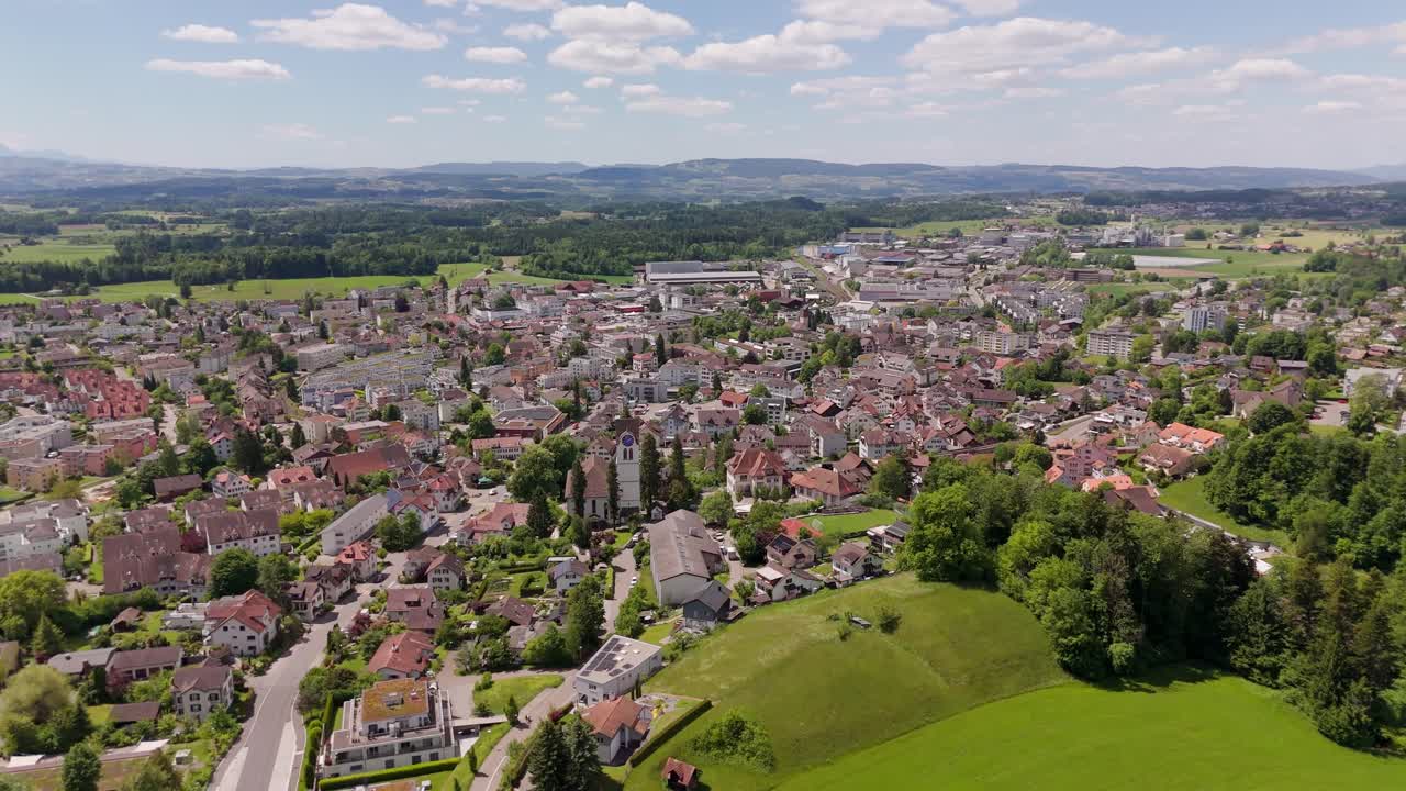 Aerial flyover historic town of Hinwil in Switzerland. Church tower and houses in center of city. Wide shot. Sunny day with green picturesque landscape in distance. Clouds at sky in spring.