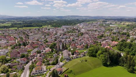 aerial flyover historic town of hinwil in switzerland. church tower and houses in center of city. wide shot. sunny day with green picturesque landscape in distance. clouds at sky in spring.