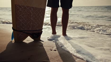 Close-up-shot-of-a-guy-in-shorts-standing-on-the-seashore-and-holding-a-surfboard-near-him-and-small-waves-passing-through-his-legs-and-leaving-sea-foam-on-them-in-the-morning-at-Sunrise