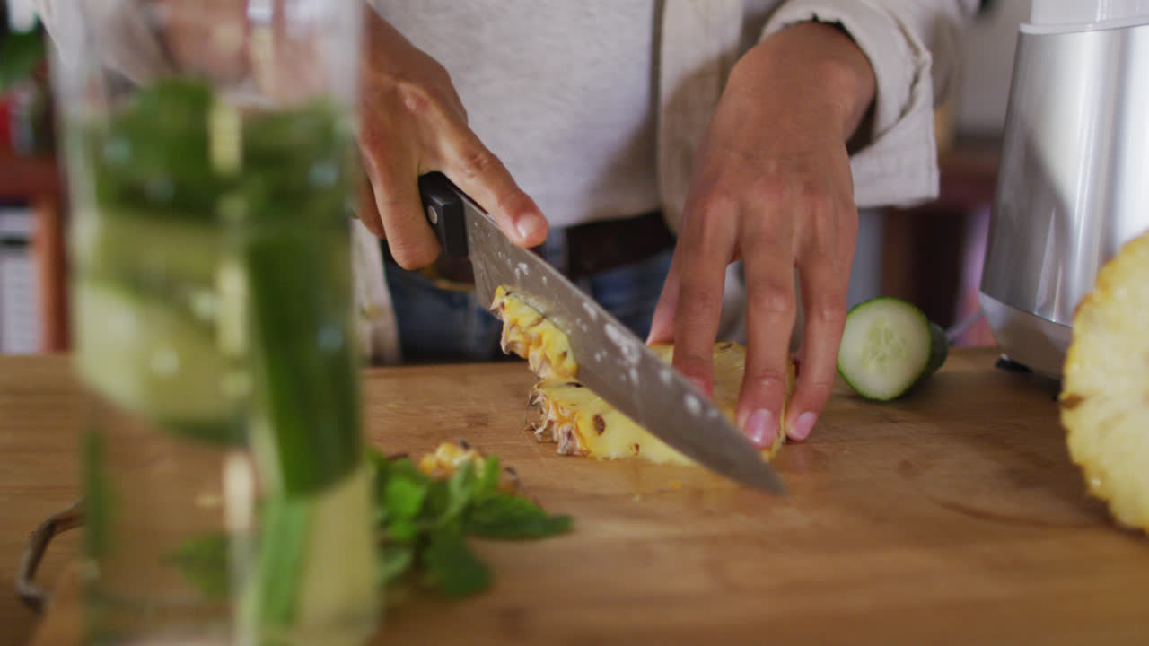 sección media de raza mixta mujer preparando bebida saludable cortando piña en la cocina de la cabaña