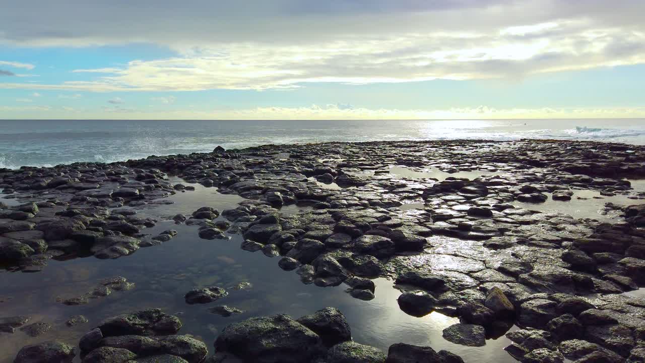 4K Hawaii Kauai boom down and left from backlit ocean waves in distance past pools on lava rock to low shot of lava rock with partly cloudy sky