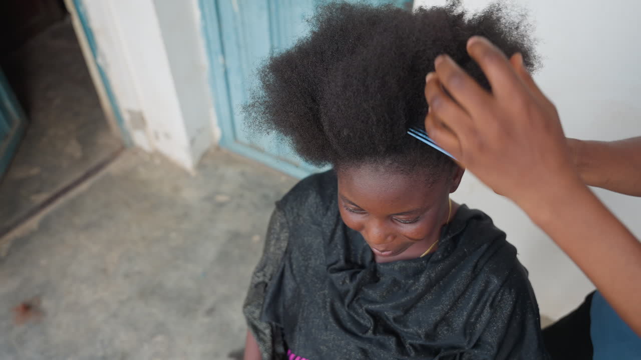 Donna di colore che si fa pettinare i capelli sull'uscio di casa con luce naturale, una parrucchiera vicina districa delicatamente l'afro vicino a una porta blu, sorriso giocoso e abbigliamento casual, mani che modellano la texture e dividono i capelli, pavimento in cemento.