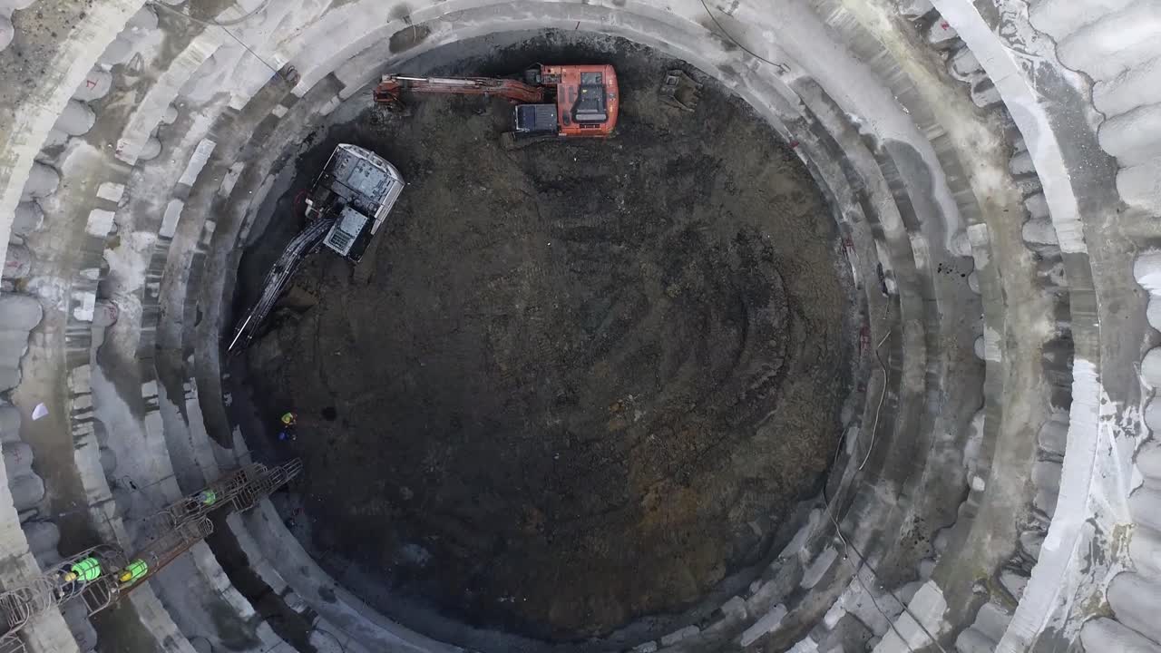 Workers climb up a lattice ladder while excavators excavate in deep tunnel shaft construction.