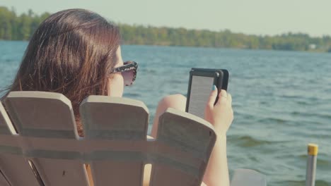 mujer joven relajándose y leyendo en una silla adirondack en un muelle con un lago en el fondo
