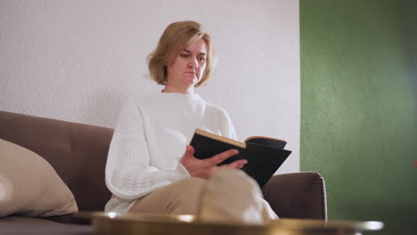 open heated woman seated on couch with legs crossed, attentively flipping through pages of book in peaceful room setting, absorbed in thoughtful reading and quiet personal reflection