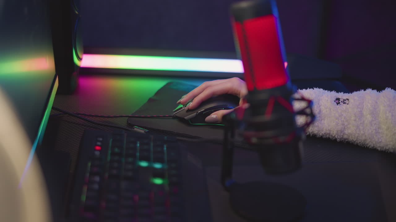 Hand view of lady with polished nail in fluffy sweater with teddy bear design using wired gaming mouse on black pad, ambient RGB lighting surrounds workspace with visible microphone