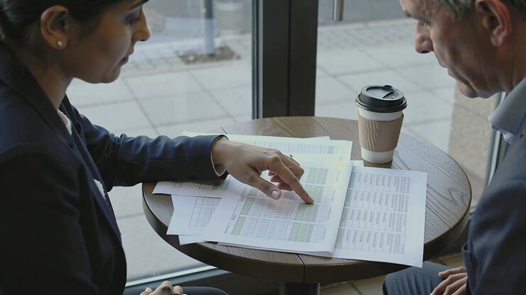 Placing hand, pointing woman in blazer guiding partner through sheets for review at cafe with cup