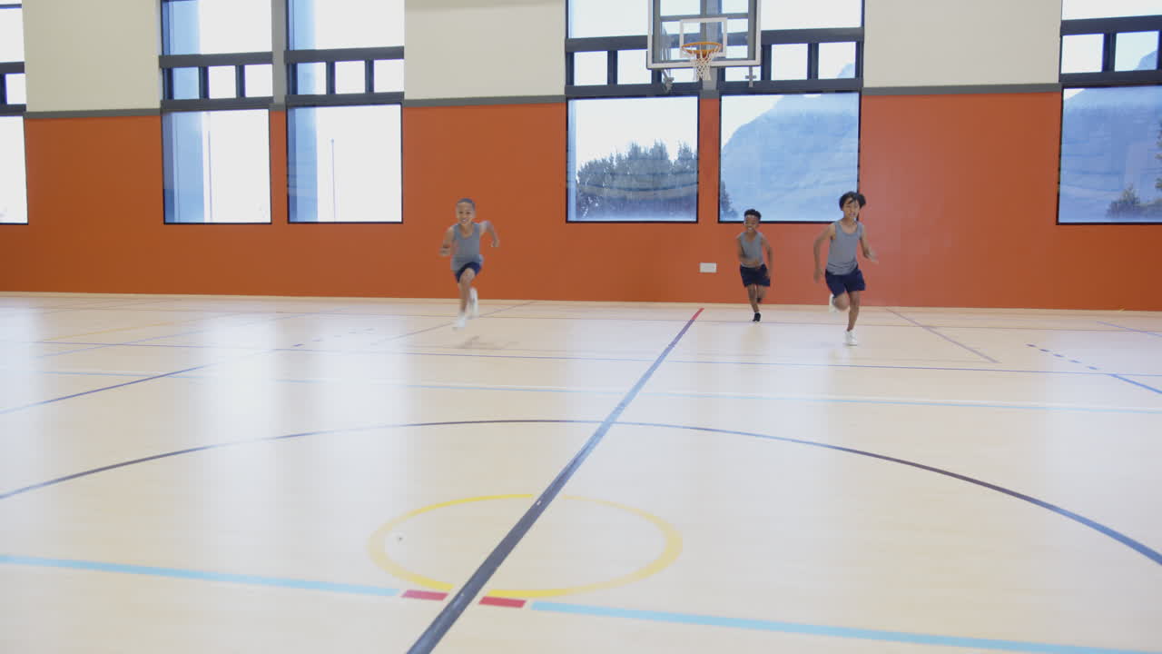 Drie jongens spelen basketbal in schoolgymzaal, rennen en dribbelen met de bal.