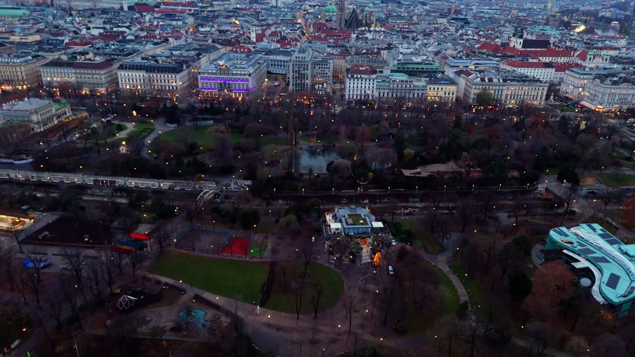 Ripresa aerea laterale di un paesaggio ripreso con drone che sorvola lo Stadtpark di Vienna in inverno, mostrando alberi spogli, sentieri del parco, prati aperti ed edifici circostanti della città sotto una tenue luce invernale