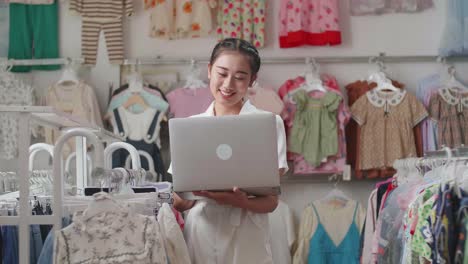 la propietaria asiática de una tienda de ropa para niños disfruta usando una computadora portátil, visitando las redes sociales y haciendo marketing en línea después de comprobar el stock en los rieles.