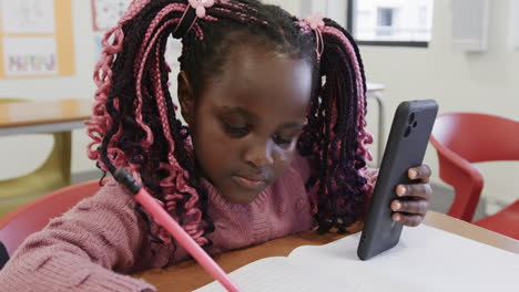 in classroom, young girl writing in notebook while holding smartphone attentively, at school