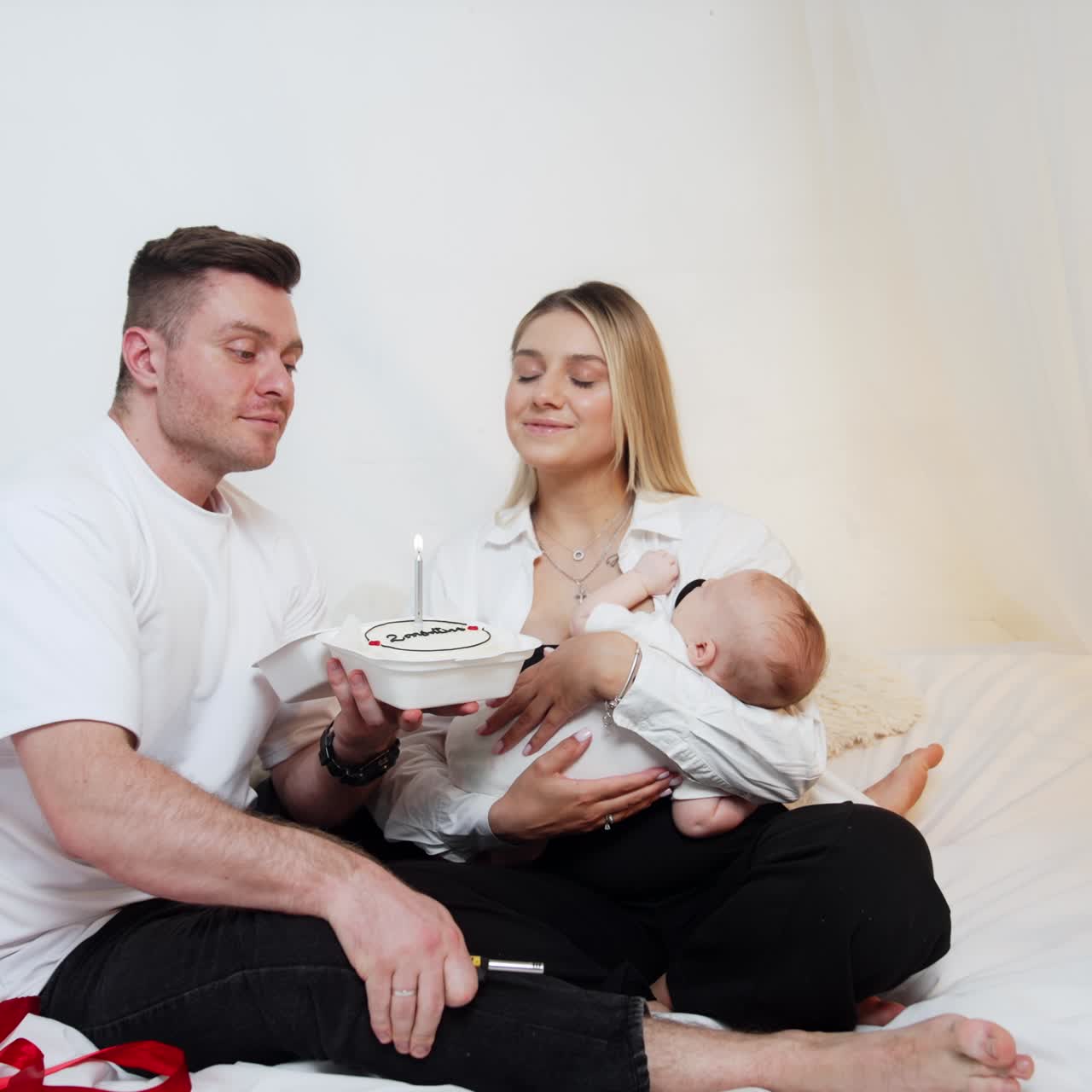 Lovely couple with a baby sits on the canopy bed. Man holds a cake and lights the candle. Parents blow the candle together making a wish