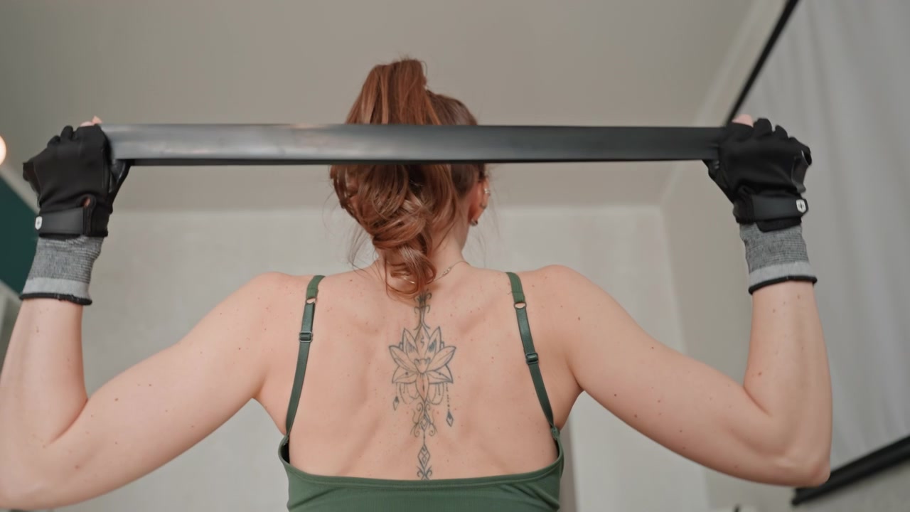 Mulher caucasiana em aquecimento fitness em casa, vista traseira mostrando elevações de ombro no tapete, regata verde, tatuagem floral nas costas, luz natural suave, ambiente de sala de estar, postura focada, rotina de mobilidade para a parte superior do corpo.