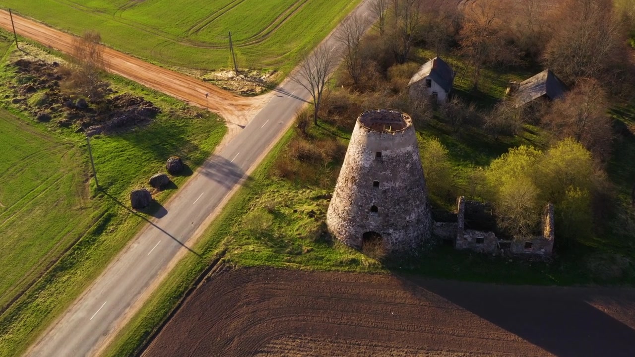 Carrellata aerea lungo un campo agricolo con inclinazione verso il basso su una struttura in mattoni di un mulino a vento in rovina