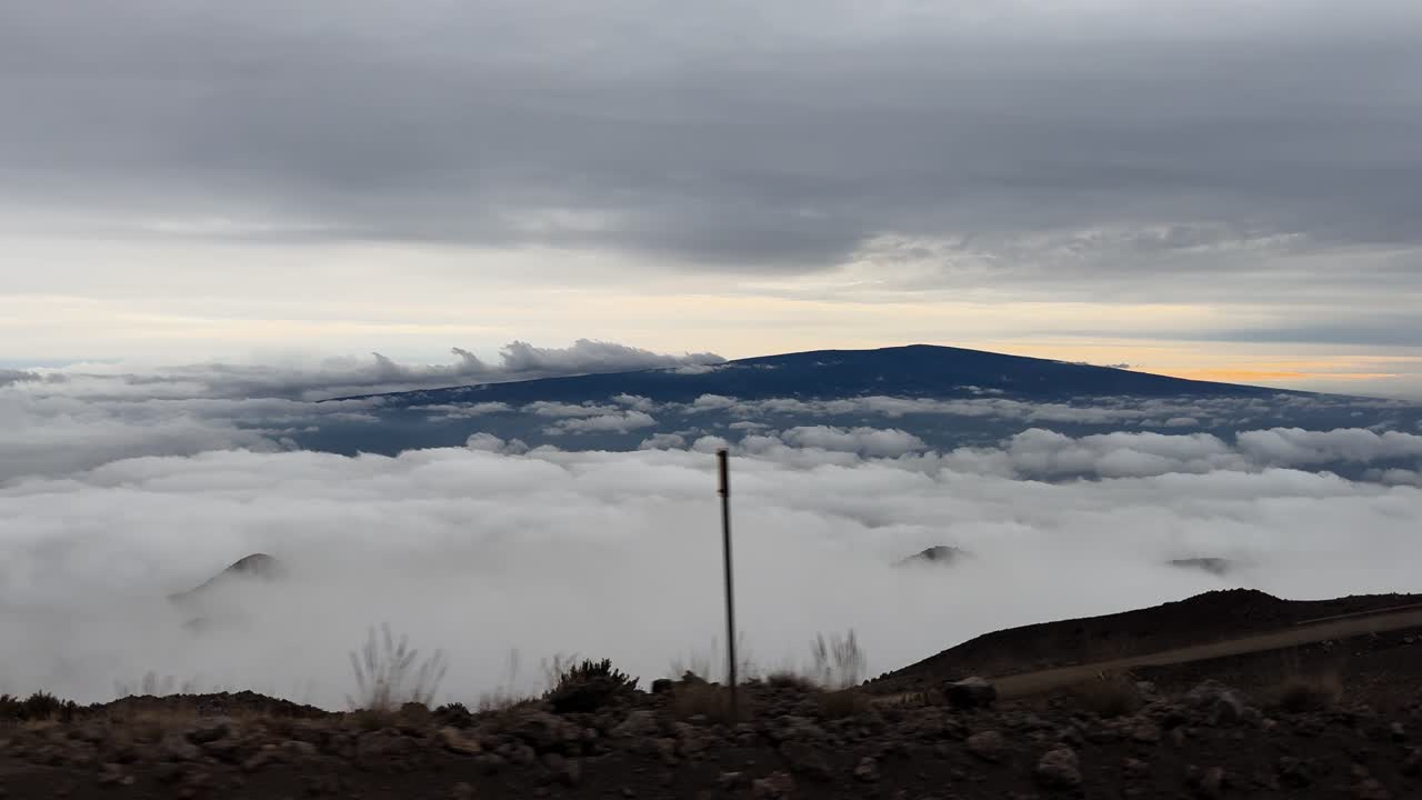 Vídeo grabado desde un vehículo en movimiento en la isla de Hawái, que muestra una densa capa de nubes cubriendo el terreno inferior, con las crestas distantes de Mauna Loa visibles sobre las nubes bajo un cielo nublado