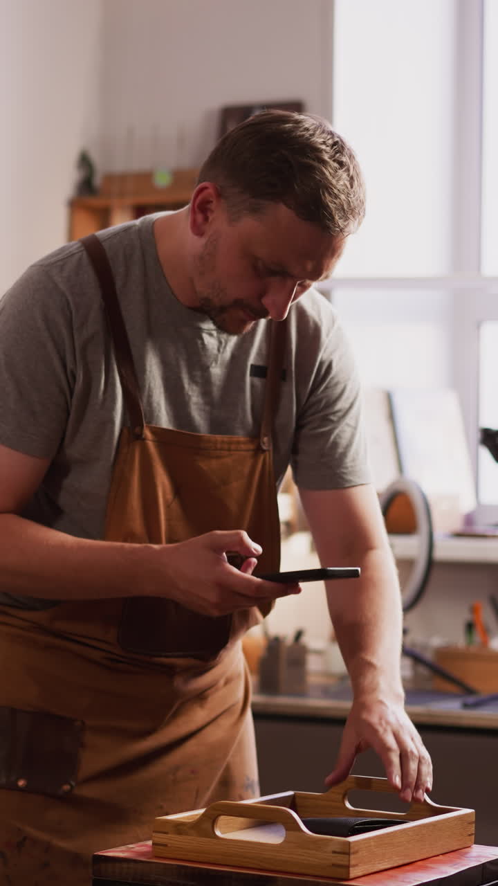 Master in apron takes picture of leather item on wooden tray with smartphone in workshop slow motion. Craftsman prepares product for sale