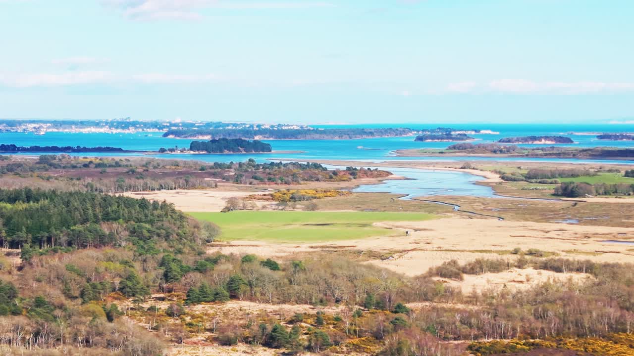 Vidsträckt vy över en kustnära flodmynning med sandbankar, slingrande vattenkanaler och små öar omgivna av landsbygd under klar himmel i Storbritannien.