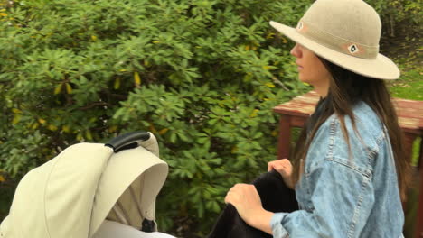side view of young woman wearing a wide-brimmed hat and denim jacket, pushing a beige baby stroller across a grassy park, capturing motherhood, outdoor activity, and family life