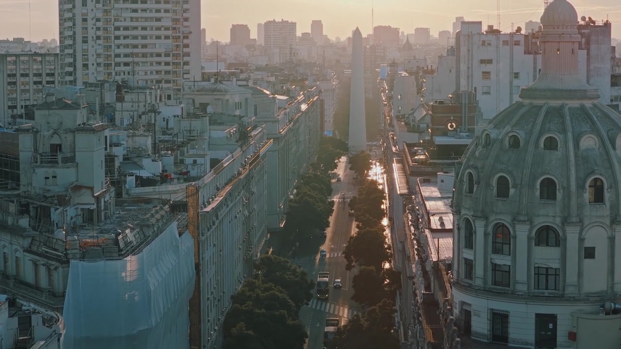 Ripresa aerea con drone che si inclina verso l'alto dal traffico stradale e dai tetti storici per rivelare l'iconico Obelisco e lo skyline al tramonto lungo viale Diagonal Norte a Buenos Aires, Argentina
