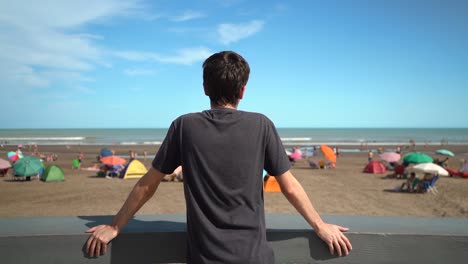 Back-View-Of-A-Young-Man-Standing-And-Watching-View-Of-Ocean-With-People-Resting-On-Tent-And-Beach-Parasol