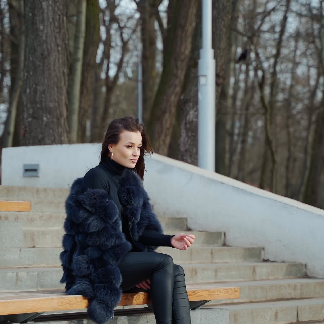 Beautiful woman sitting outdoors on the background of leafless trees. Side view of a sexy woman in black fur coat resting in the park. Slow motion.