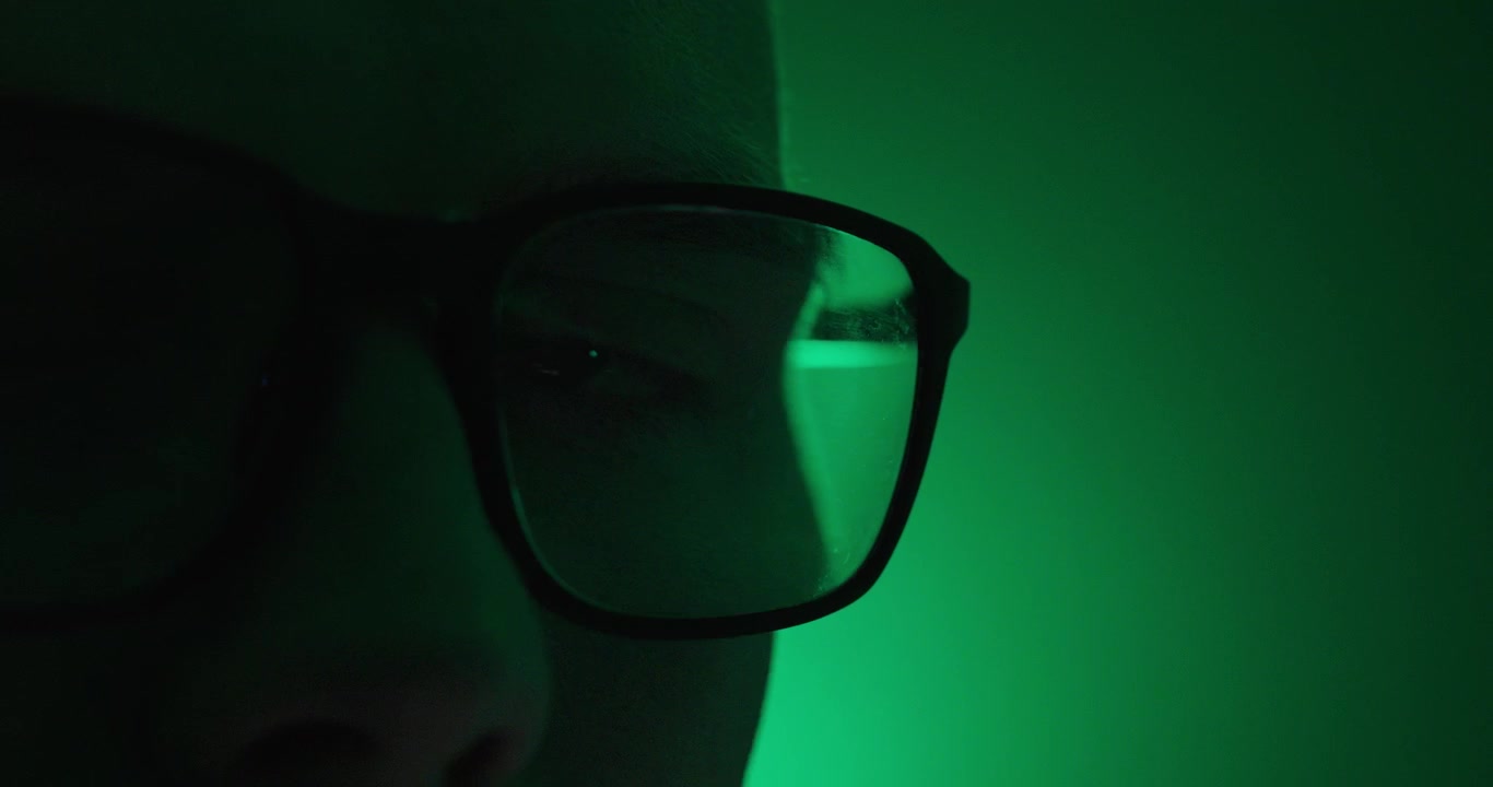 close-up of a person's eye behind eyeglasses illuminated by dramatic green neon and strobe lighting. Technology, cybersecurity, focus, mystery, and futuristic portrait concept.
