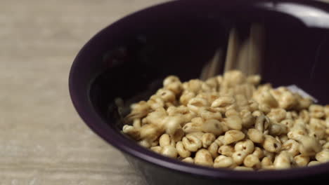 Close-up-of-honey-wheat-cereal-falling-into-empty-purple-bowl,-profile