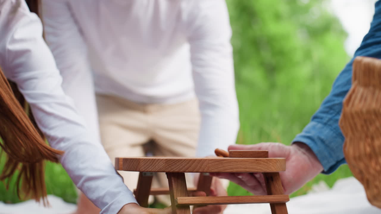 키 큰 녹색 나무와 밝은 햇빛으로 둘러싸여 따뜻한 가족 피크닉 분위기가 연출되는 야외에서 아이들이 아버지를 도와 아버지가 작은 나무 테이블을 피크닉 매트 위에 조심스럽게 놓는 모습을 낮은 각도에서 보여줘요.