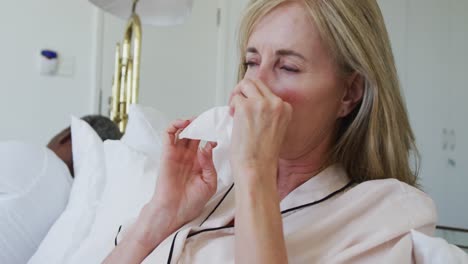 diverse senior couple in bed woman blowing nose and coughing while man sleeps