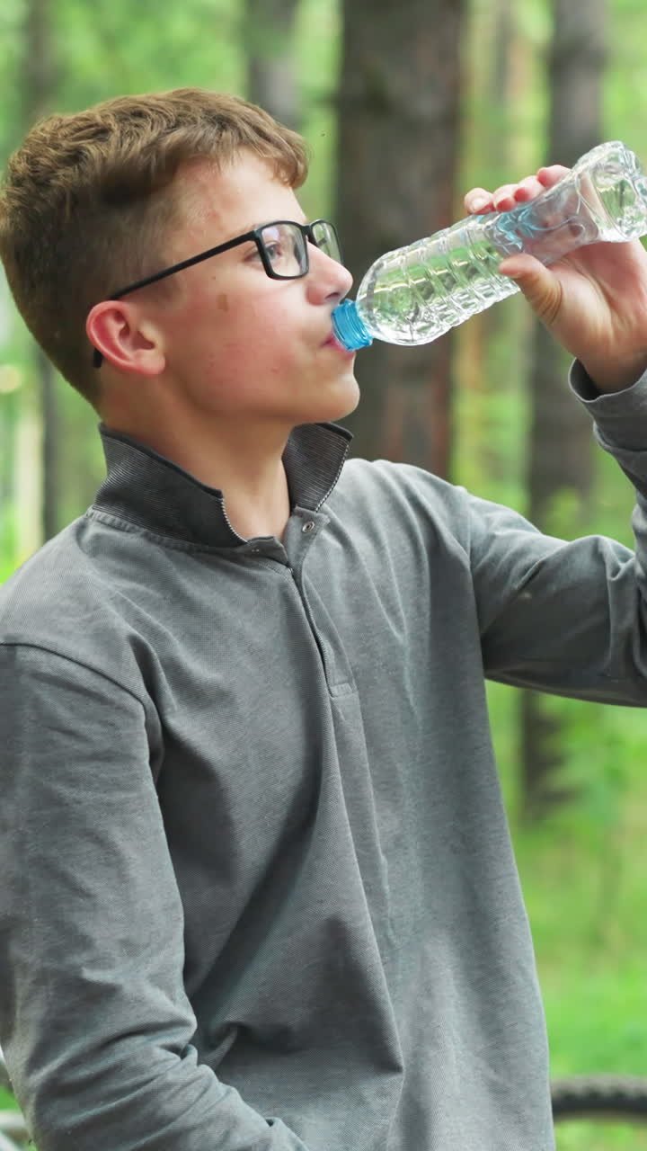 안경을 쓰고 회색 장갑 을 입은 어린 소년은 물 한 병을 열고 숲에서 휴식을 취하는 동안 한 잔을 마시며, 키가 큰 녹색 나무로 둘러싸인 배경에 자전거가 주차되어 있습니다.