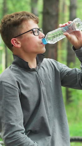 un niño joven con gafas y una camiseta de manga larga gris abre una botella de agua toma un sorbo, mientras toma un descanso en un bosque, la bicicleta está estacionada en el fondo, rodeada de altos árboles verdes