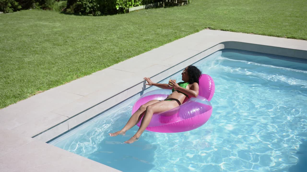 Donna afroamericana in bikini su un galleggiante rosa nella piscina di casa con una bevanda, che afferra la sponda per stabilizzare il galleggiante