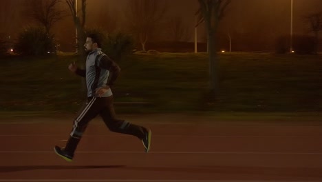 Video-En-Cámara-Lenta-De-Un-Joven-Corriendo-En-Una-Pista-De-Carreras-Por-La-Noche-Mientras-Usa-Ropa-Deportiva