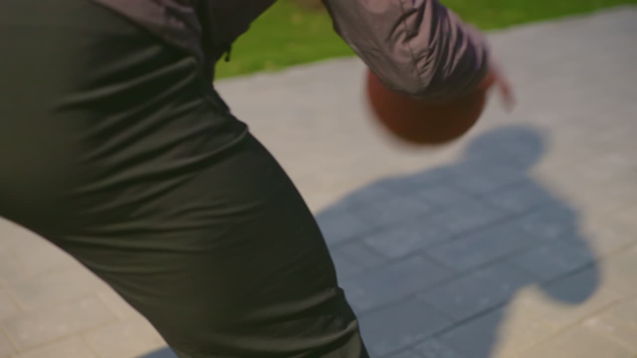 Closeup af dribling på solbeskinnet fortov, streetball-scene i midtbyen med spiller, der laver crossover, orange basketball der hopper tæt på lang skygge, sorte joggingbukser og mørk jakke, lavt hoftestilling, hurtig