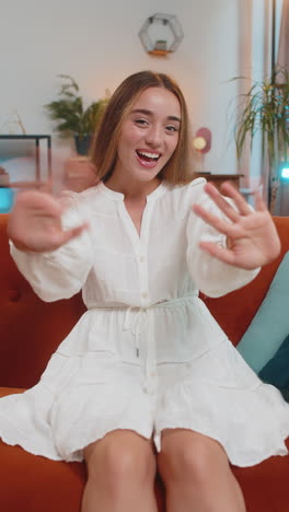 happy young woman vlogger smiling friendly at camera and waving hands while sitting on couch at home