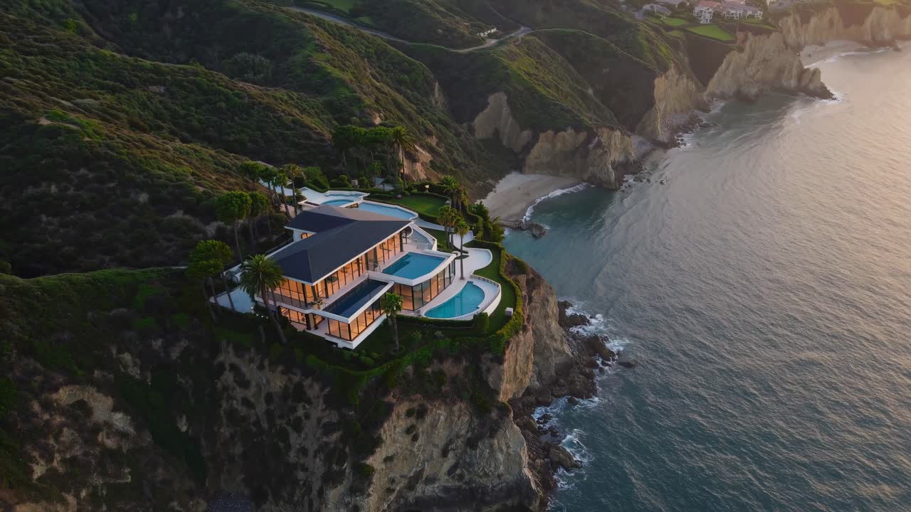 Modern glass and concrete home sitting on a cliff overlooking the ocean at sunset, featuring a swimming pool and lush landscaping, offering breathtaking views of the California coastline