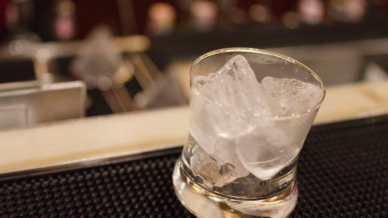 Bartender drops ice cubes into glass at luxury bar, warm lighting, shallow depth of field